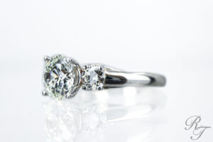 A custom engagement ring featuring a large round brilliant diamond center stone flanked by a round diamond side stone, set in a polished white gold three-stone setting, handcrafted by Rick Terry Jewelry Designs in Knoxville, TN.
