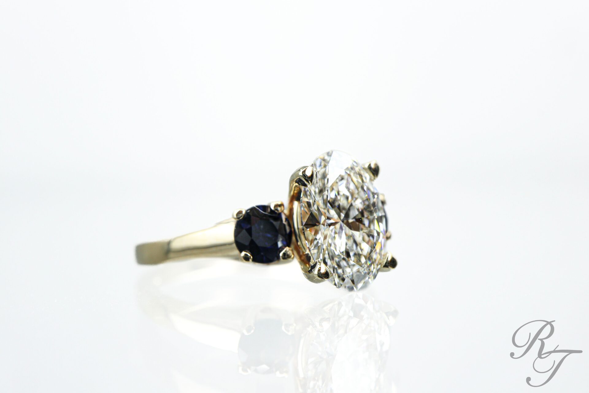 Side profile of an oval diamond engagement ring with a six-prong setting and blue sapphire side stones, illustrating healthy, secure prongs from Rick Terry Jewelry Designs.